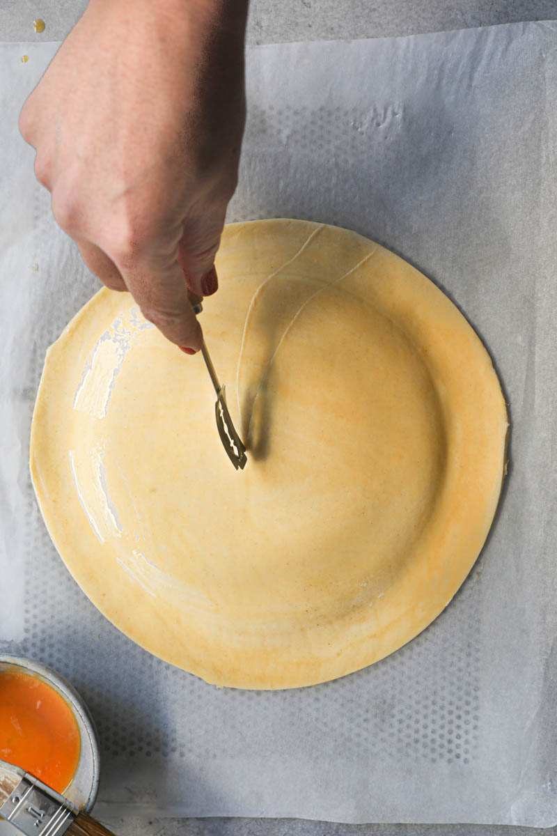 One hand scoring the French king cake with a scoring knife.