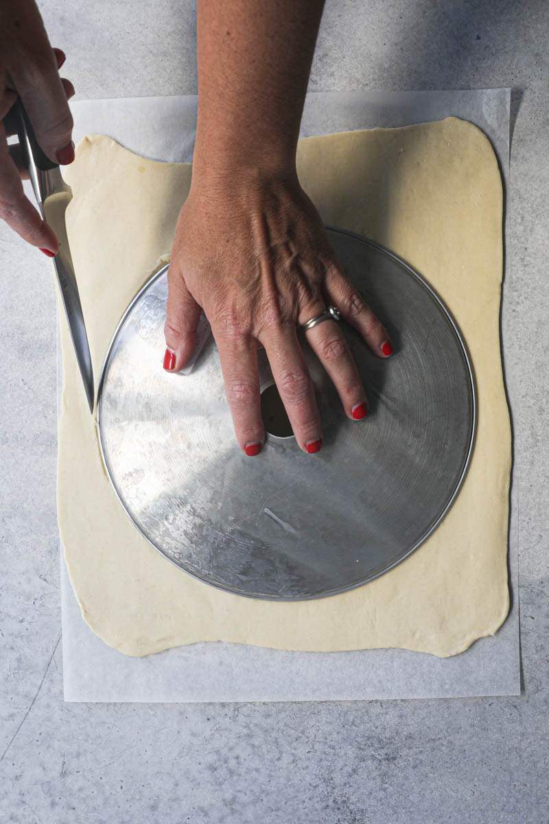 One hand holding a cutter over the puff pastry and another hand cutting around it with a knife.