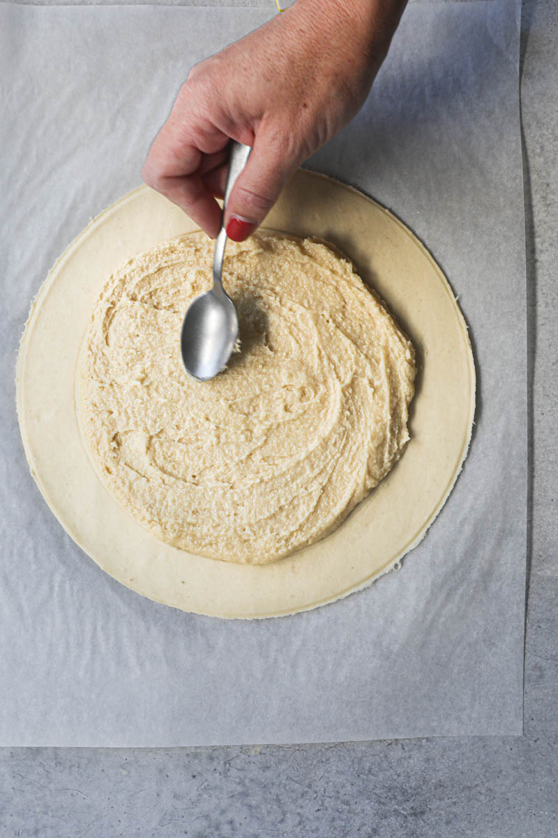 One hand spreading the almond cream over the puff pastry round with a spoon.