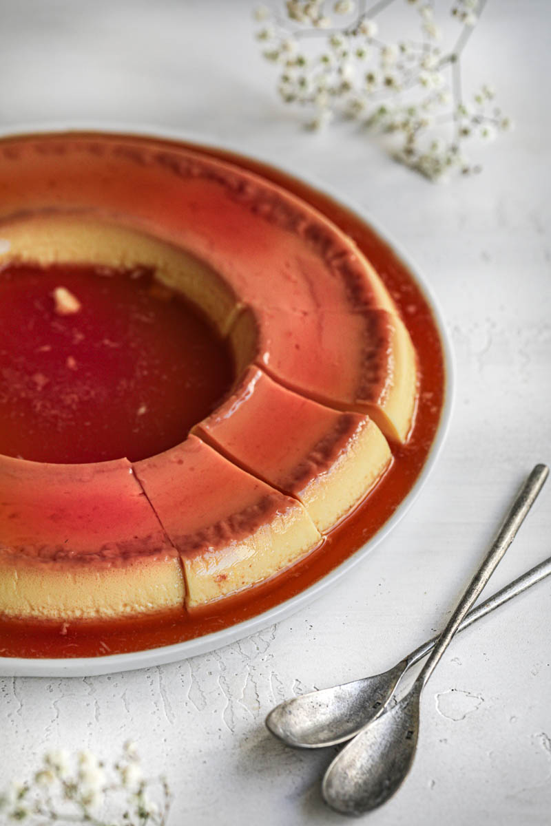 The sliced homemade crème caramel seen from the side, on a white plate with some white flowers in the back and in the left bottom corner.