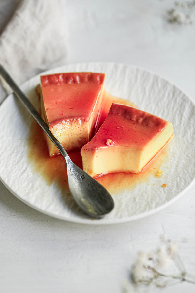 2 slices of flan on a small white plate with a silver spoon on the side.