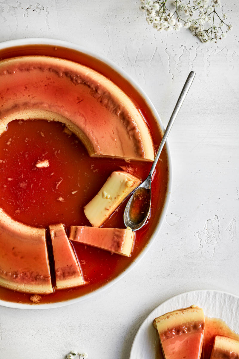 The sliced homemade flan as seen from above on a white plate, with a spoon on it, some white flowers around and a small plate with a slice of flan in the bottom right corner.