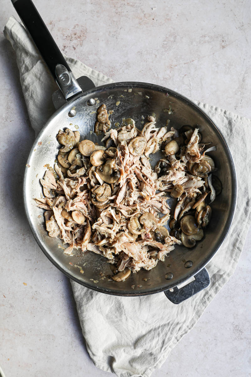 A pan on a beige linen, with the cooked shallots, the cooked sliced mushrooms, and the pulled roast chicken.