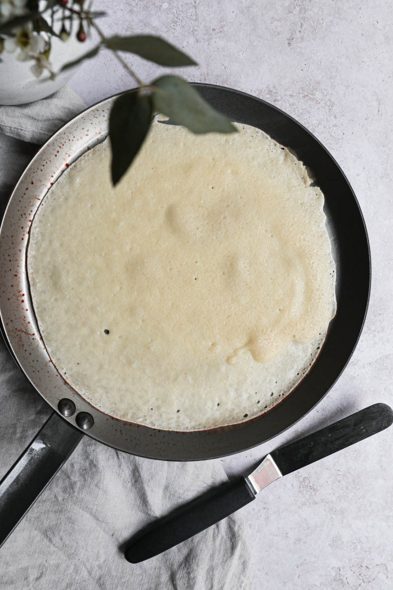 The crepe cooking in the crepe pan with a small spatula on the side.