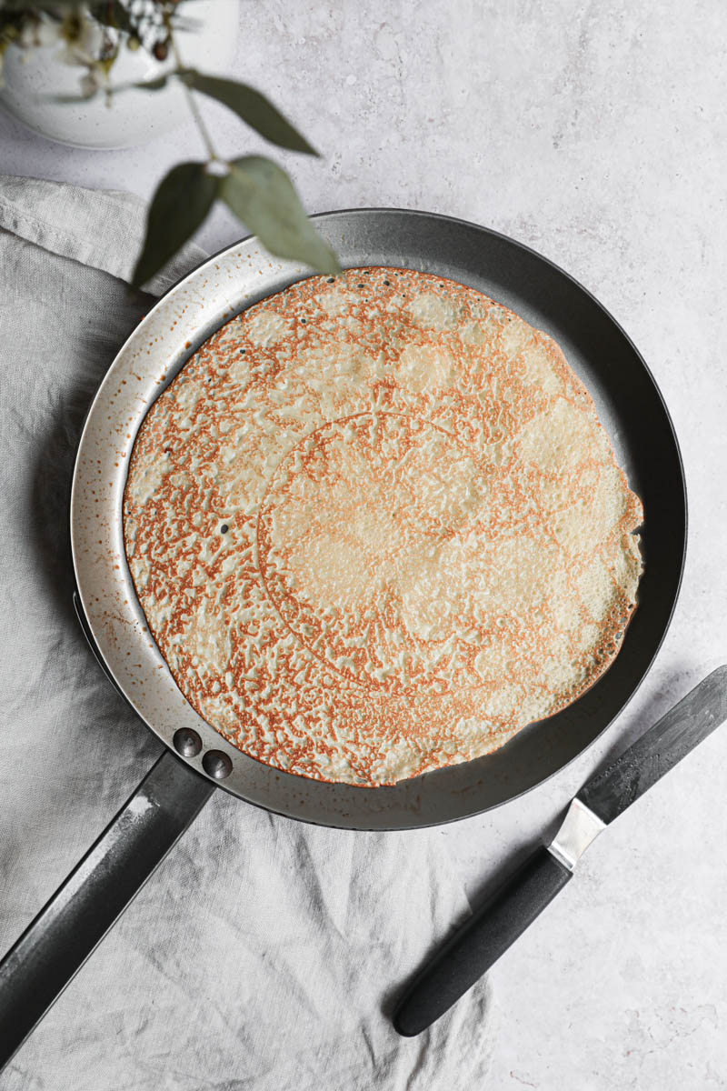 The crepe flipped in the crepe pan with a small spatula on the side.