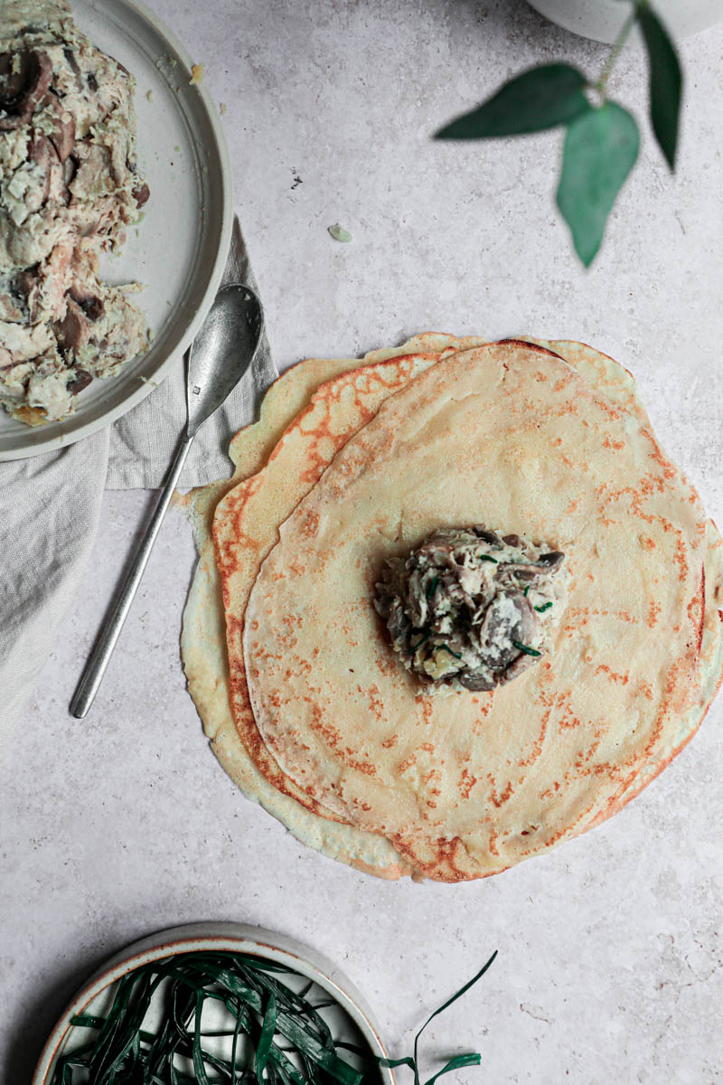 One crepe filled with a scoop of the chicken filling in the middle, with a plate filled with the filling on the top left corner and a plate with the blanched chives at the bottom.