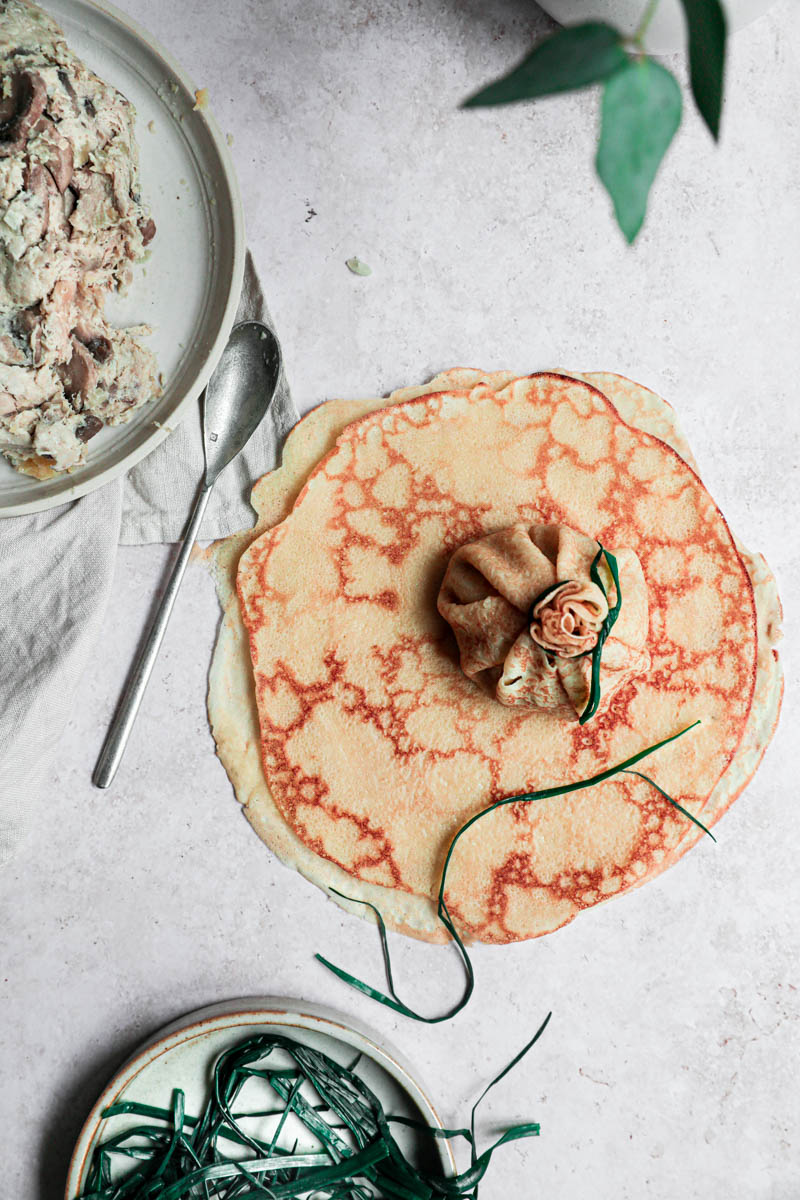 One crepe closed and tied with a chive, with a plate filled with the filling on the top left corner and a plate with the blanched chives at the bottom.