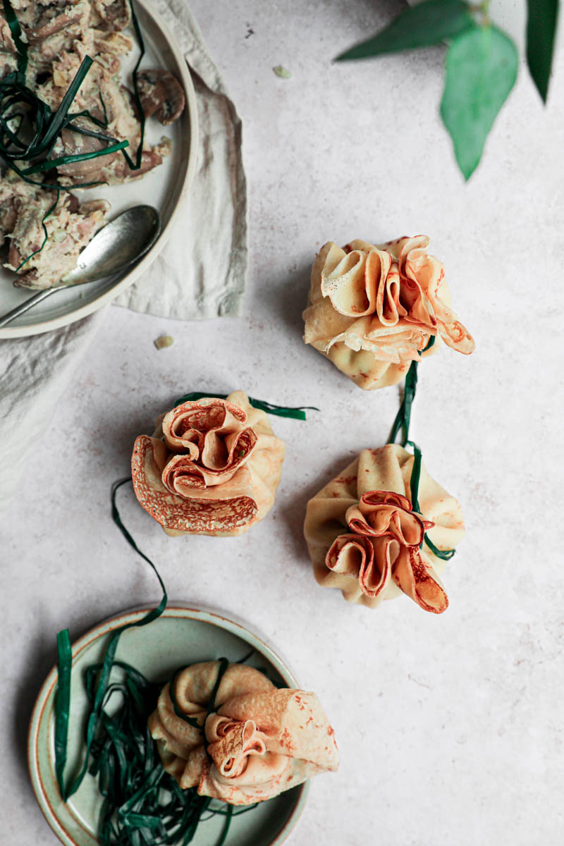 Three crepes closed and tied with a chive, with a plate filled with the filling on the top left corner and a plate with the blanched chives at the bottom.