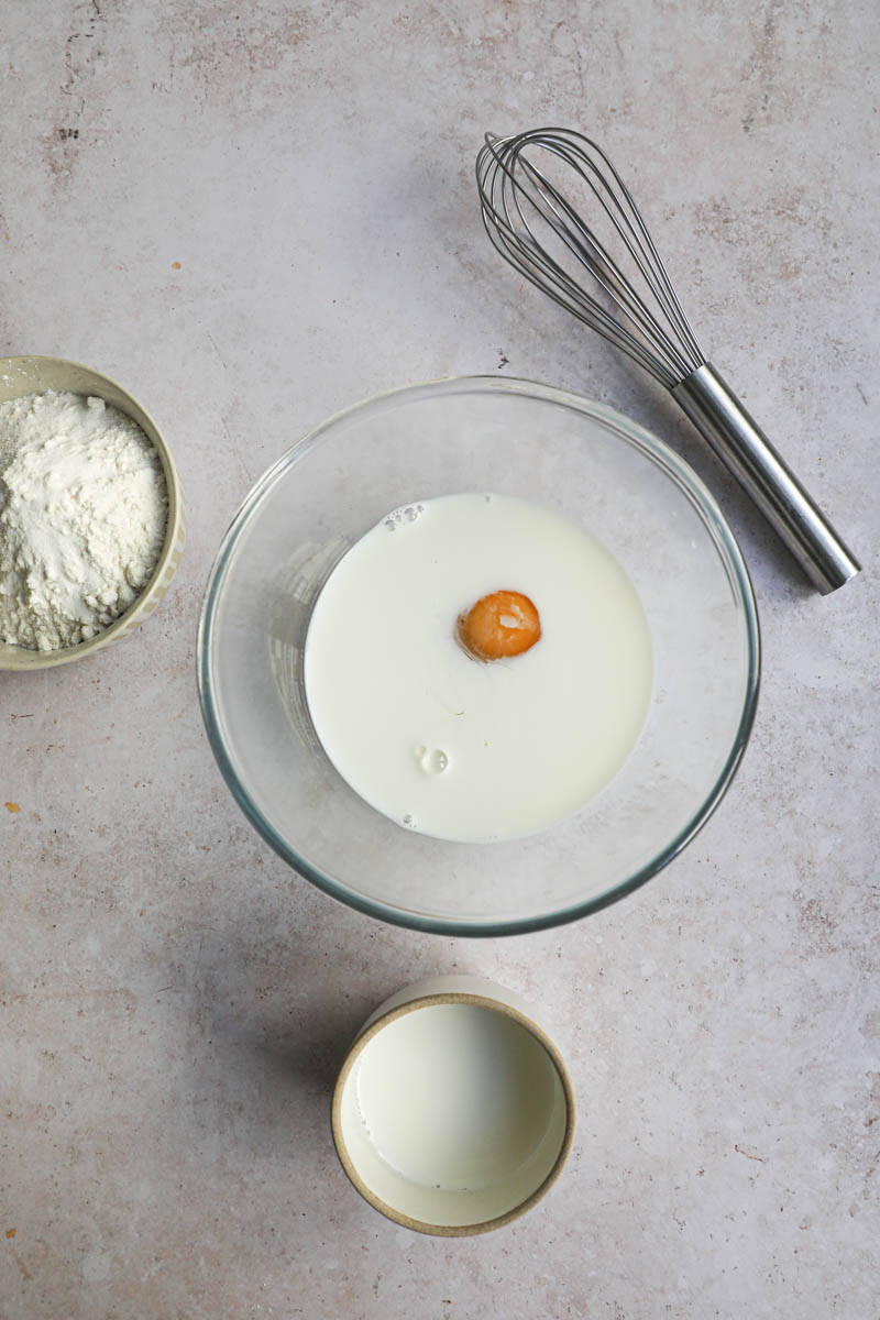 A bowl with the milk and the egg inside, with a whisk, the flour, and the butter on the side of the bowl.