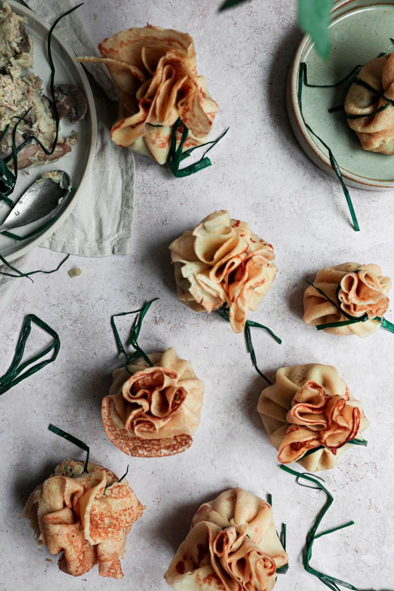 Many crepes closed and tied with a chive, with a plate filled with the filling on the top left corner and a plate with the blanched chives at the bottom.