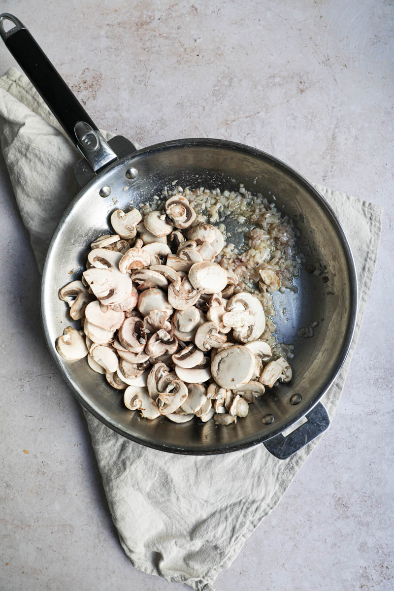 A pan on a beige linen, with the cooked shallots and the sliced mushrooms.