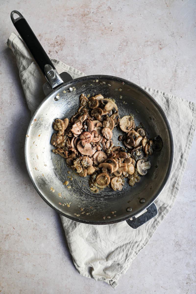 A pan on a beige linen, with the cooked shallots and the cooked sliced mushrooms.