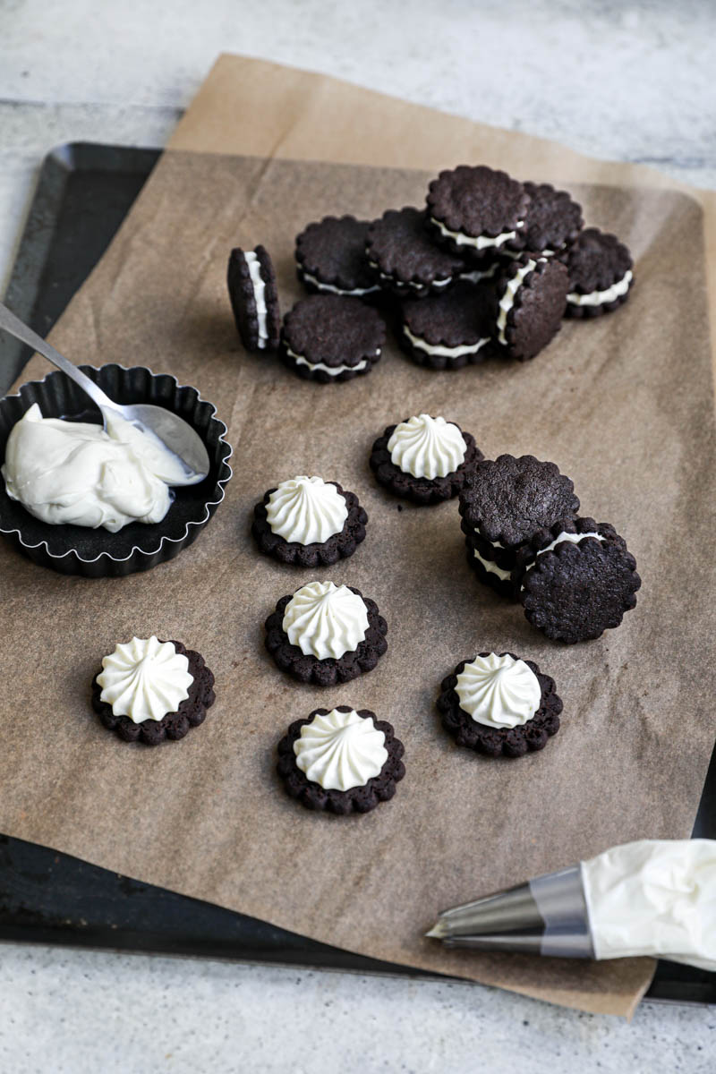 Assembly the homemade Oreo Cookies: some filled homemade Oreo cookies on top a brown piece of parchment paper, with other already filled cookies in the back, a bowl with ganache on the side and pipping bag in the front of the frame.