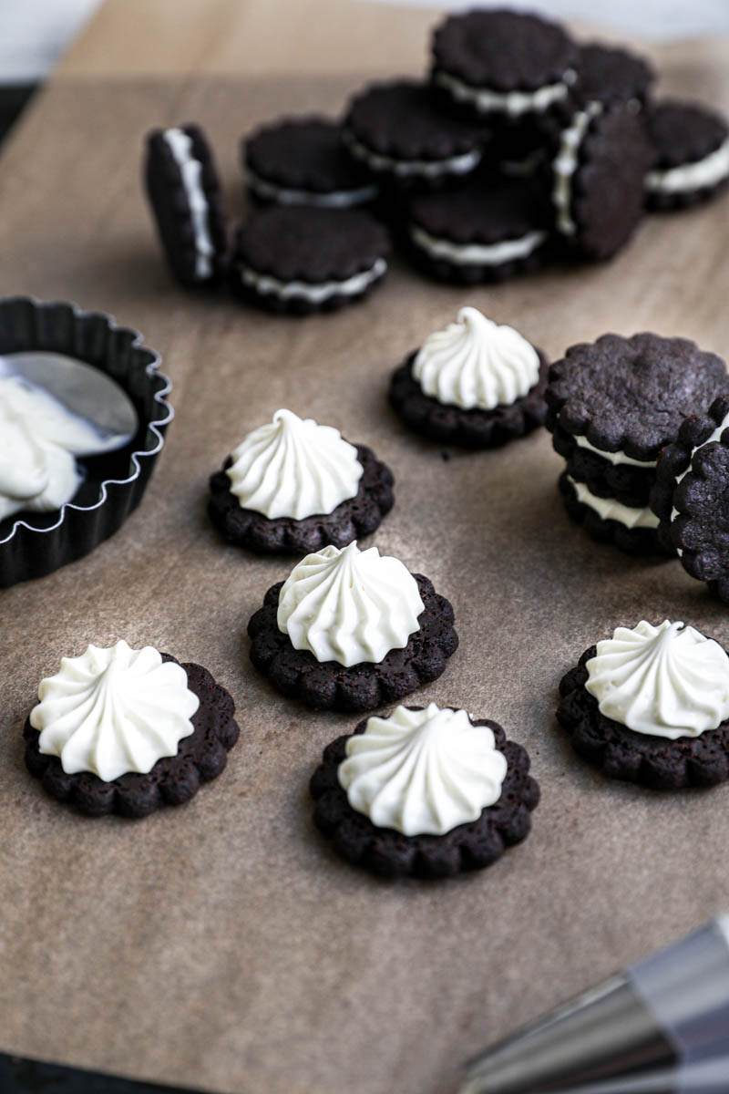 Assembly the homemade Oreo Cookies: some filled homemade Oreo cookies on top a brown piece of parchment paper, with other already filled cookies in the back, a bowl with ganache on the side and pipping bag in the front of the frame.