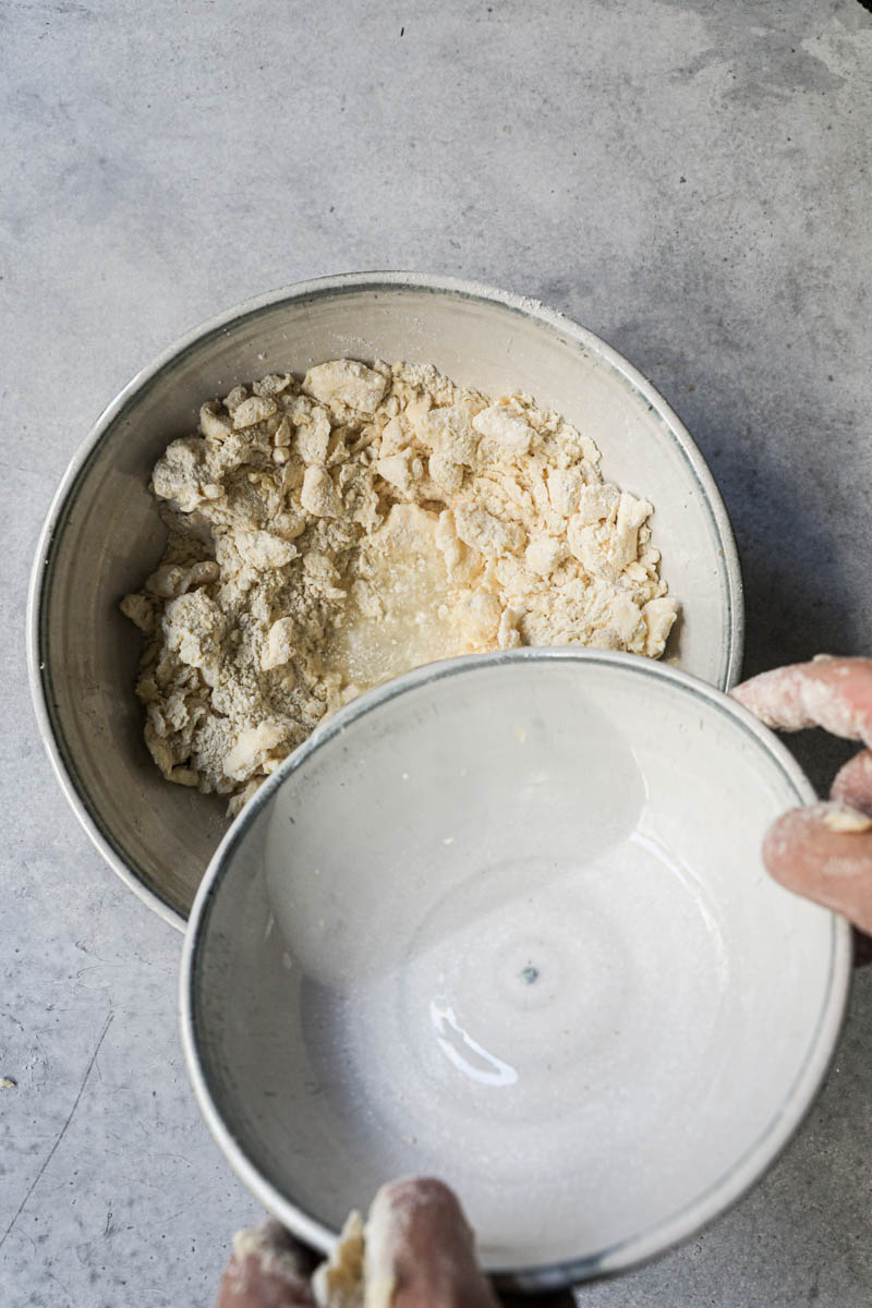 2 hands pouring water at the center of the bowl to make the rough puff pastry dough.