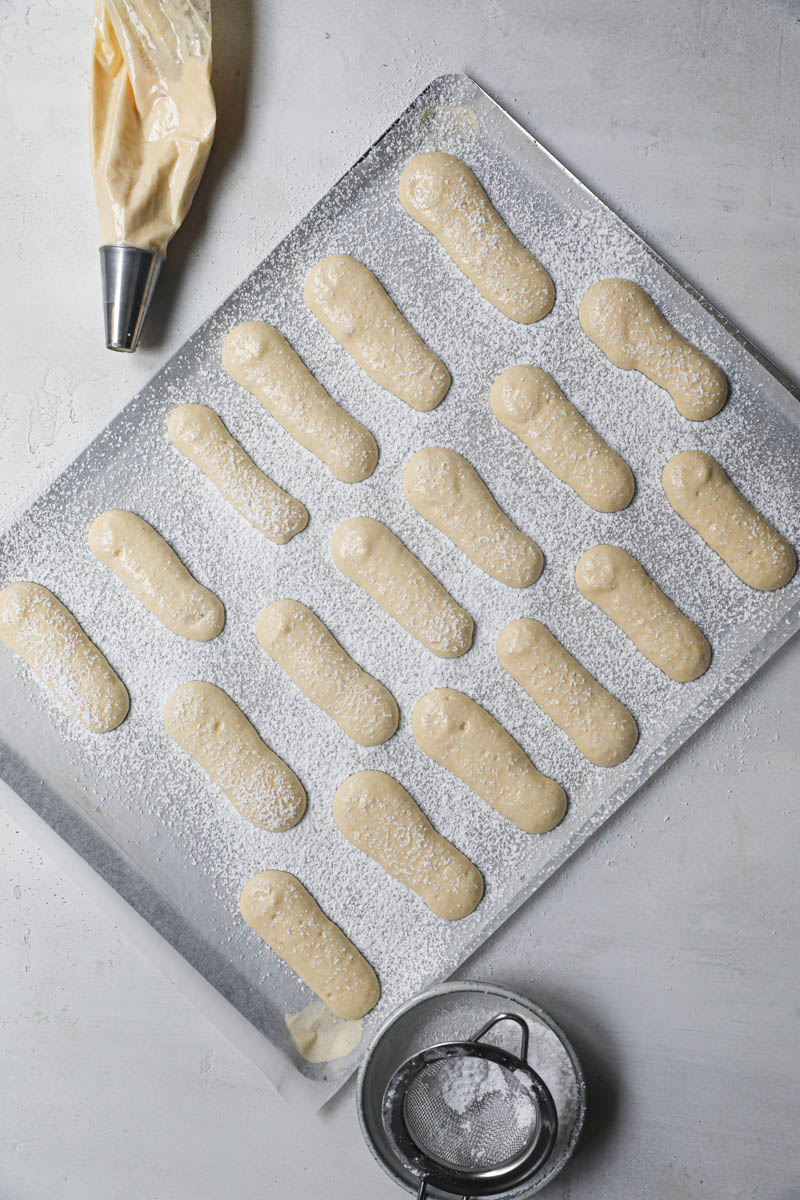 The ladyfingers cookies pipped on top of a baking tray lined with parchment paper srpinkled with powdered sugar with a piping bag in top left corner and bowl with powdered sugar in the bottom corner.