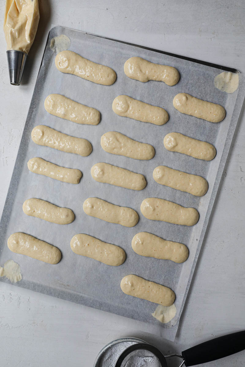 The ladyfingers cookies pipped on top of a baking tray lined with parchment paper with a piping bag in top left corner and bowl with powdered sugar in the bottom corner.