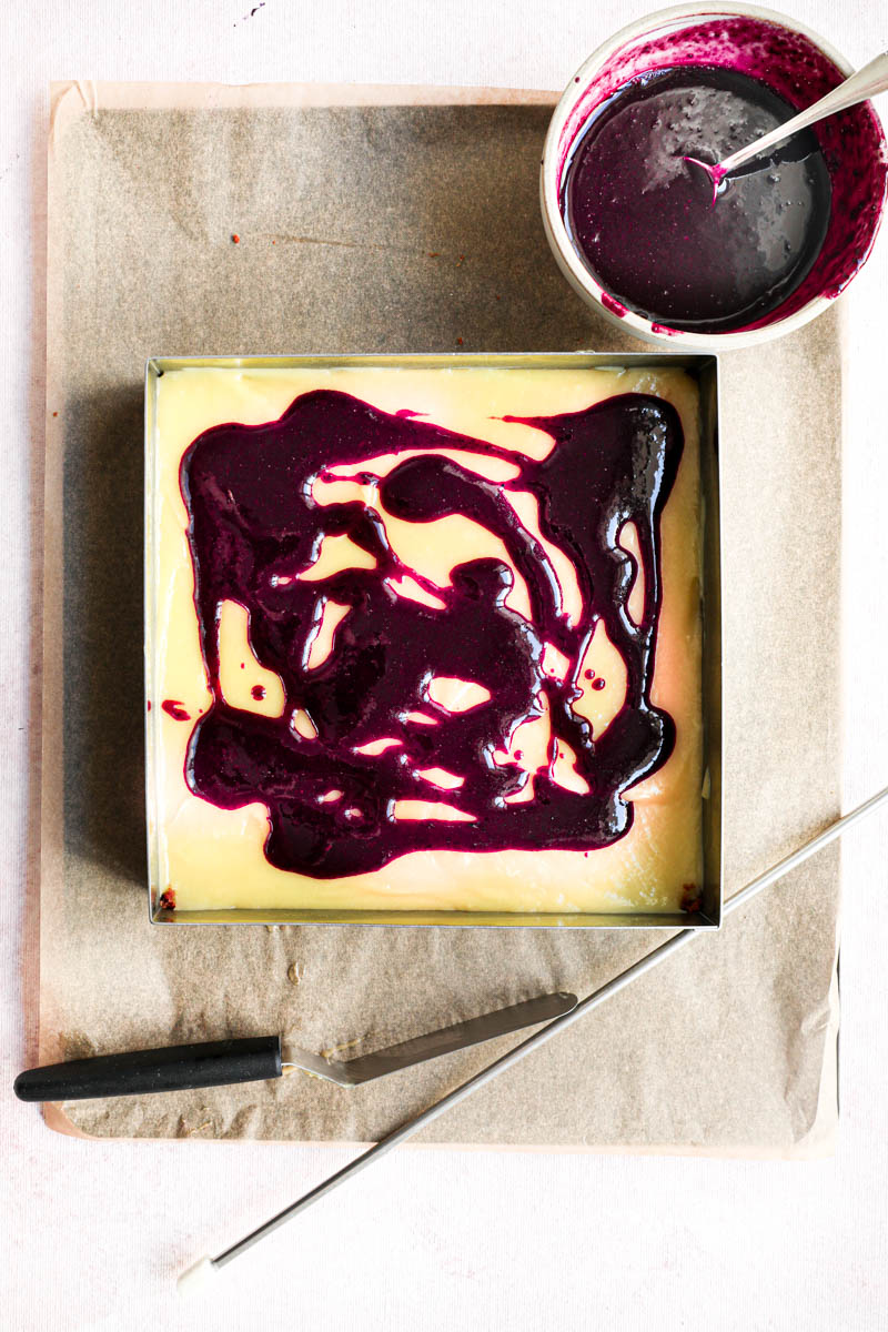 Swirling in the blueberry sauce to the blueberry lemon bars: The cake ring placed on a baking tray lined with parchment paper filled with the lemon cream, topped with blueberry sauce.