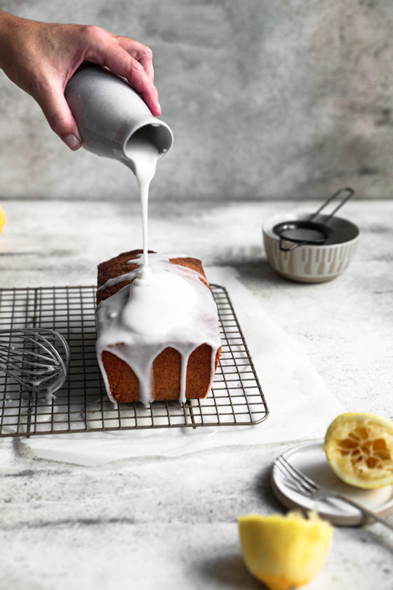 90° shot of one hand pouring the lemon glaze over the lemon cake