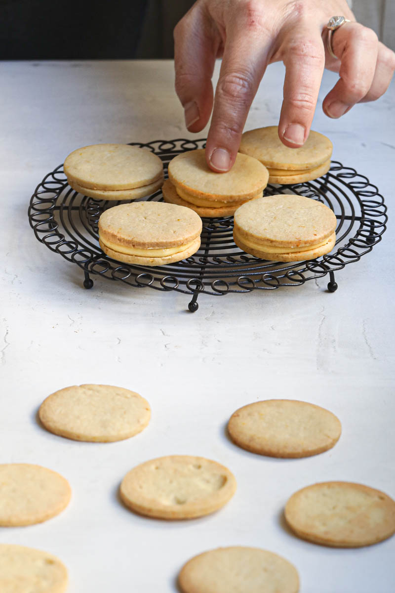 One hand placing a cookie on top of another cookie filled with lemon curd.