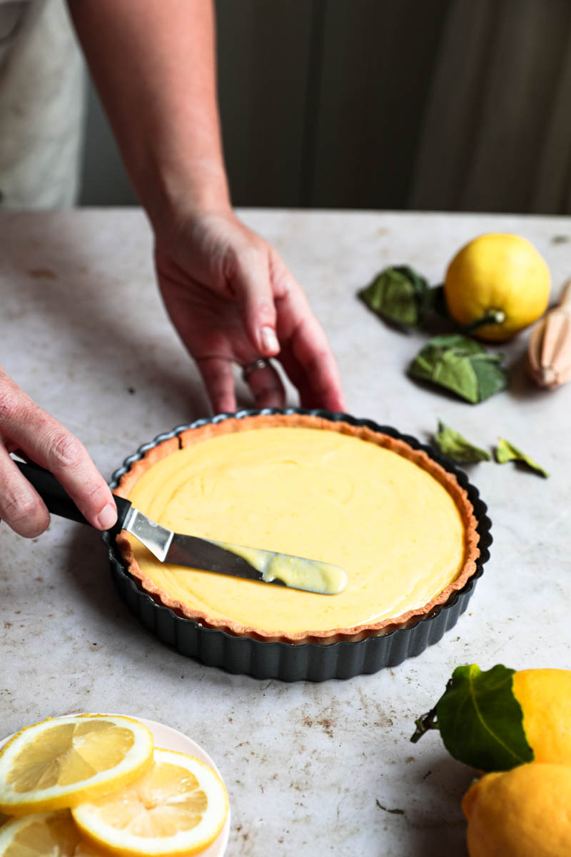 Two hands spreading the lemon curd on the tart using a small offset spatula.