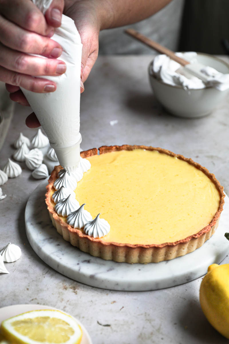 Two hands piping the swiss meringue onto the lemon curd tart.