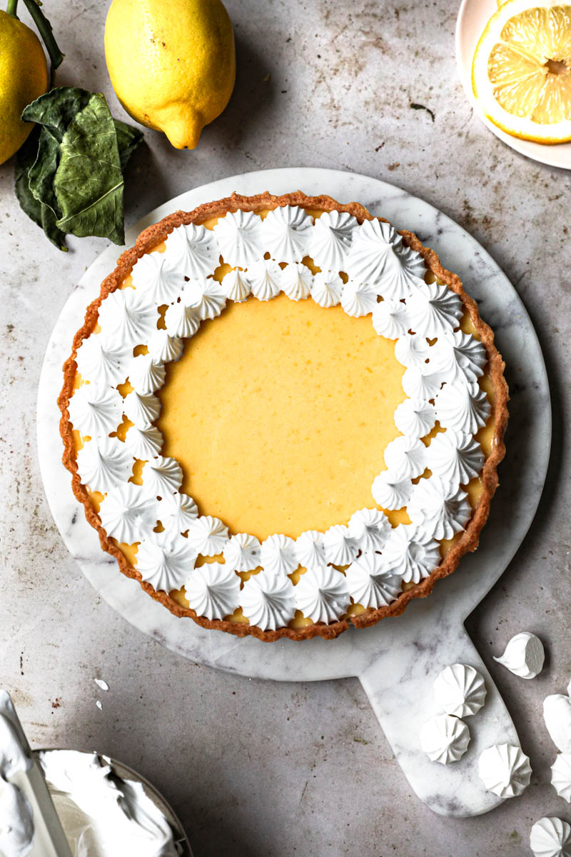The lemon curd tart topped with swiss meringue kisses with small bowl and a spatula with meringue on the side and lemon slices around.