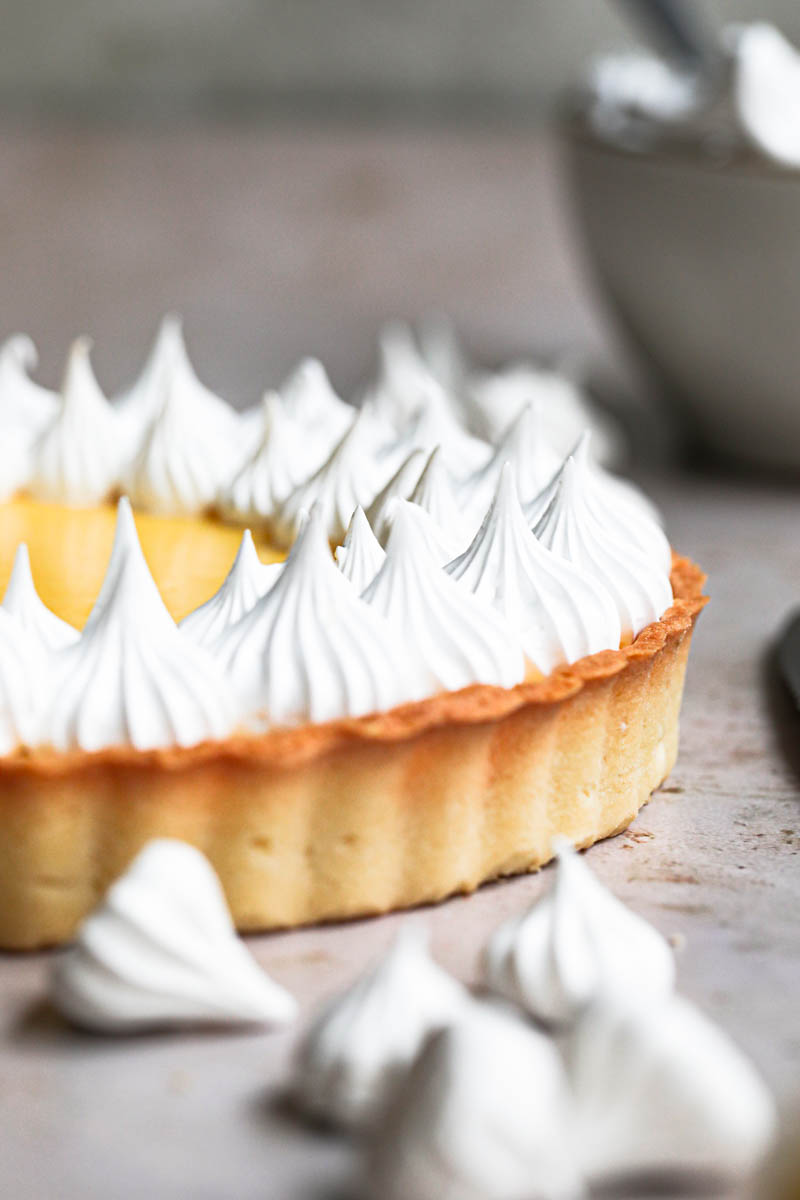 The lemon curd tart topped with swiss meringue kisses with small bowl and a spatula with meringue behind it.