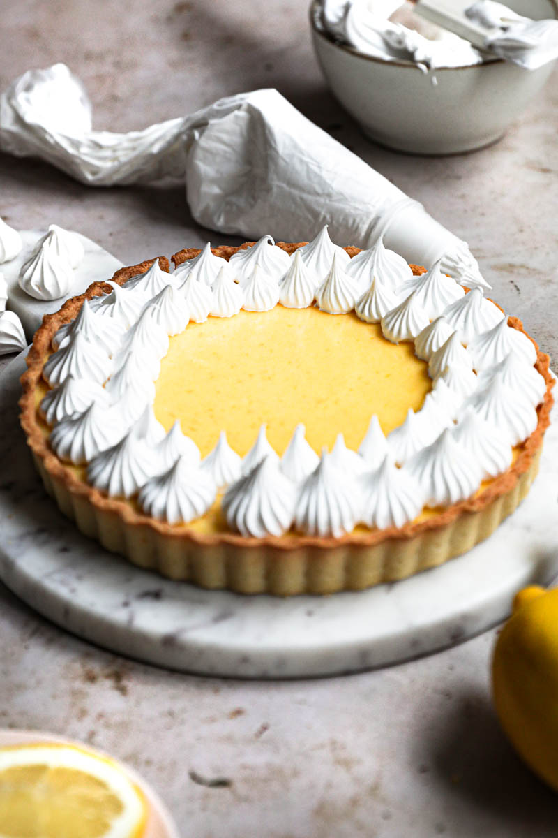 Lemon curd tart topped with swiss meringue kisses on a marble platter with a pipping bag behind the tart and some meringue kisses around.