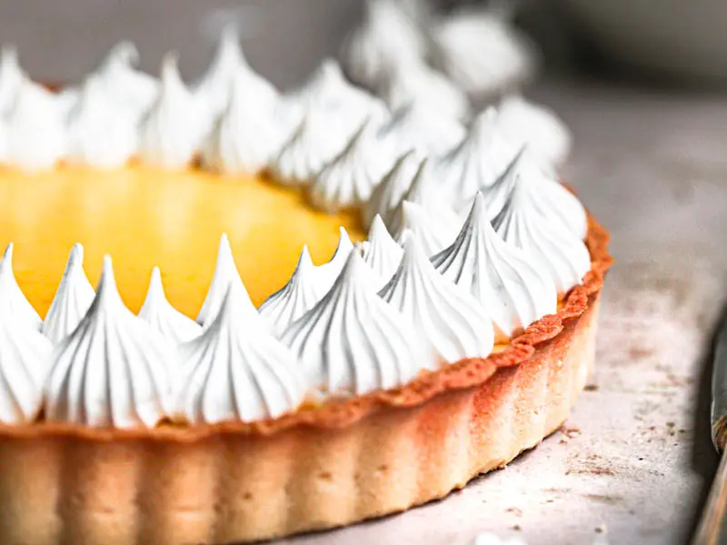 The lemon curd tart topped with swiss meringue kisses with small bowl and a spatula with meringue behind it and some meringue kisses around.