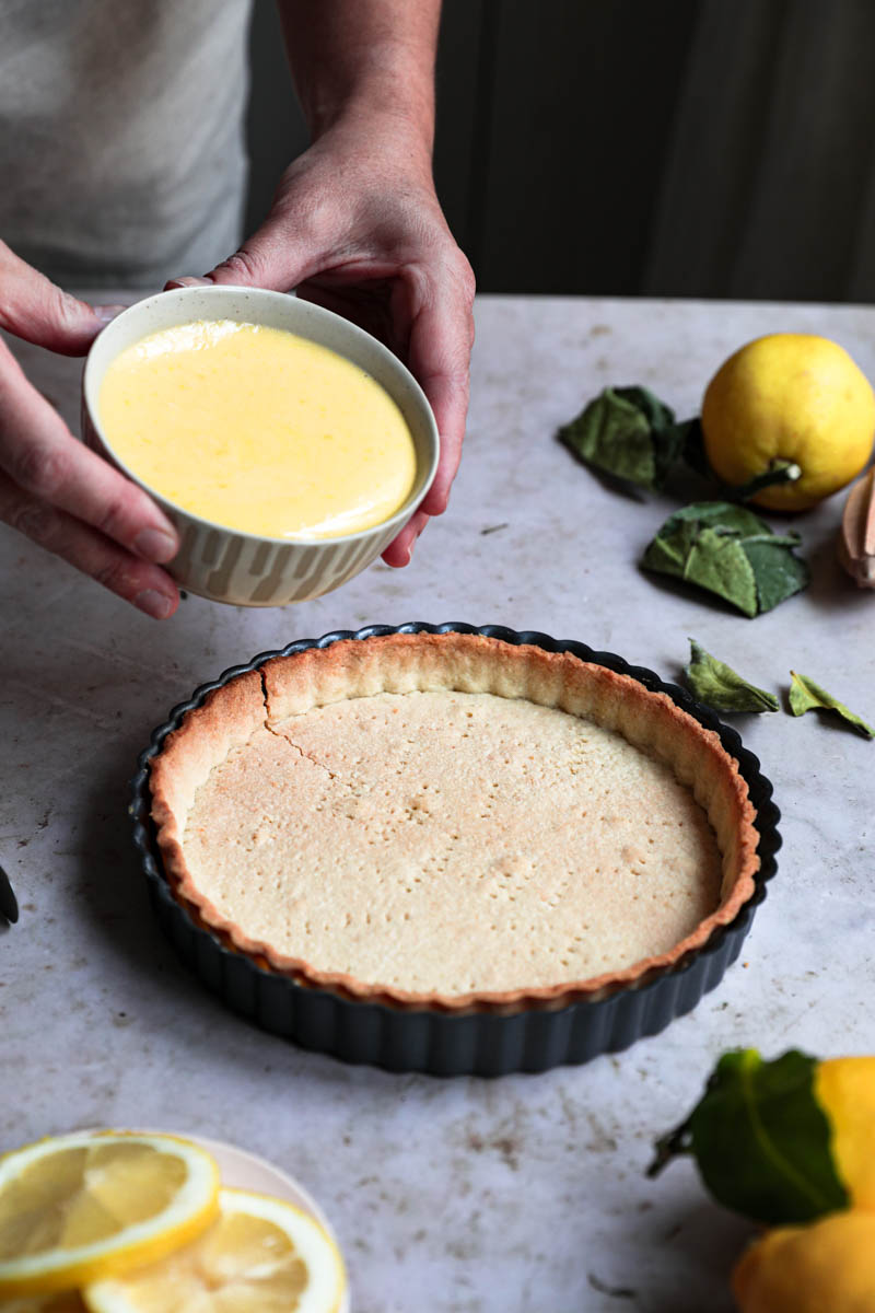 Two hands holding a small bowl with the lemon curd about to pour it over the baked tart crust.