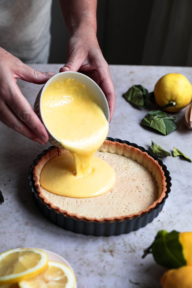 Two pouring the lemon curd about to pour it over the baked tart crust that is surrounded by lemons.