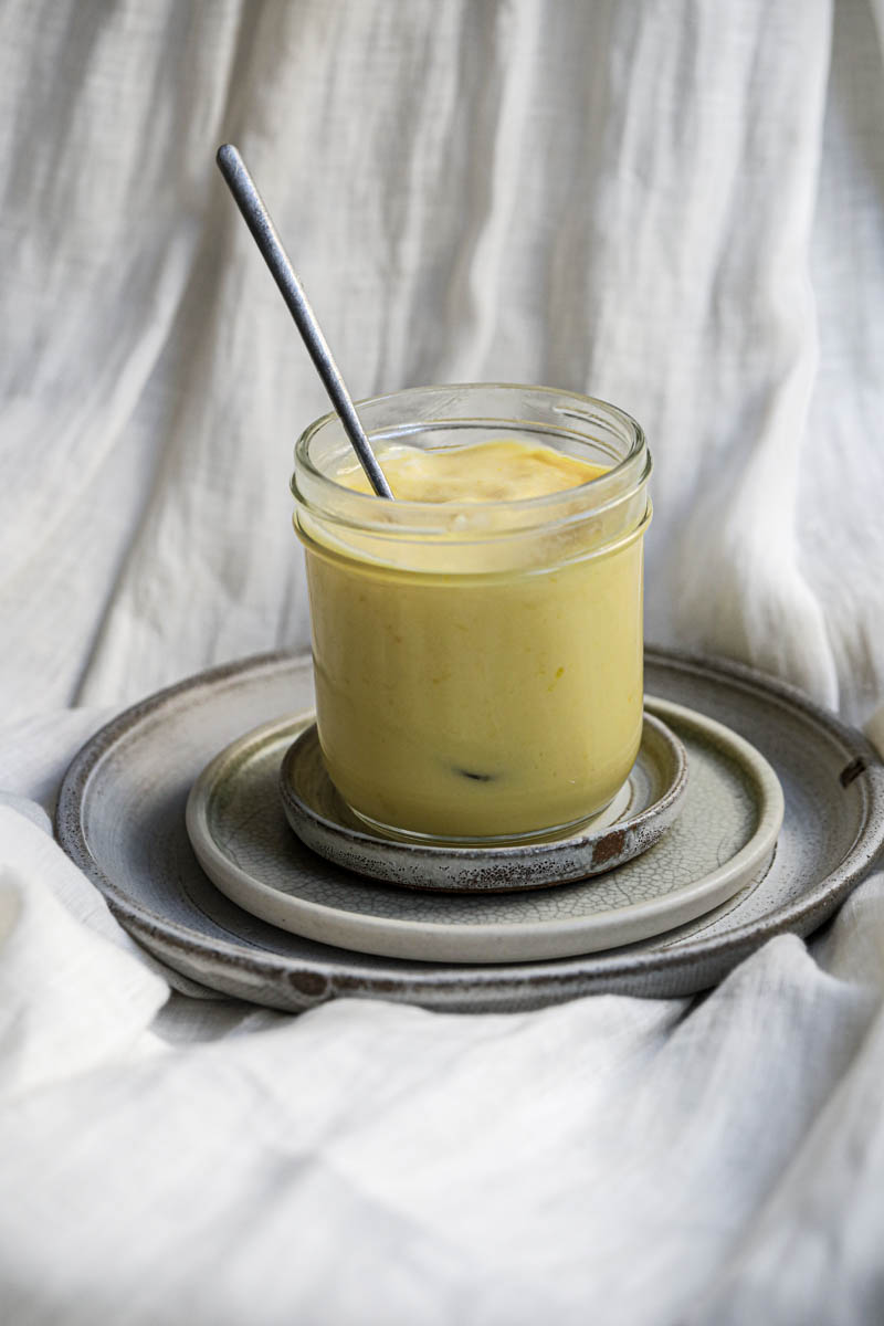 The lemon curd inside a glass jar placed on top of 3 plate with a spoon inside of the jar.
