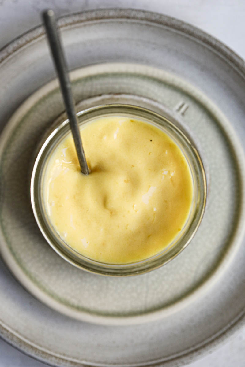 The lemon curd inside a glass jar placed on top of 3 plate with a spoon inside of the jar as seen from above.