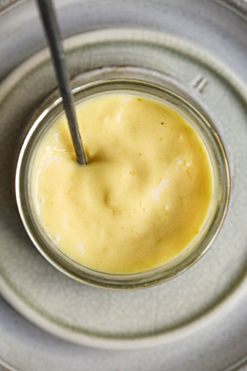Closeup of the lemon curd inside a glass jar placed on top of 3 plate with a spoon inside of the jar as seen from above.