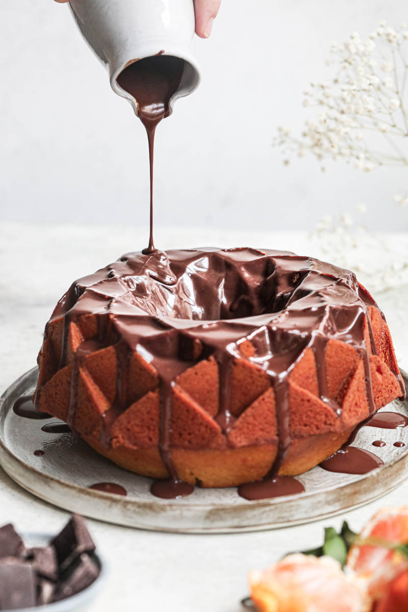 1 hand pouring chocolate ganache over the mandarin orange Bundt cake.