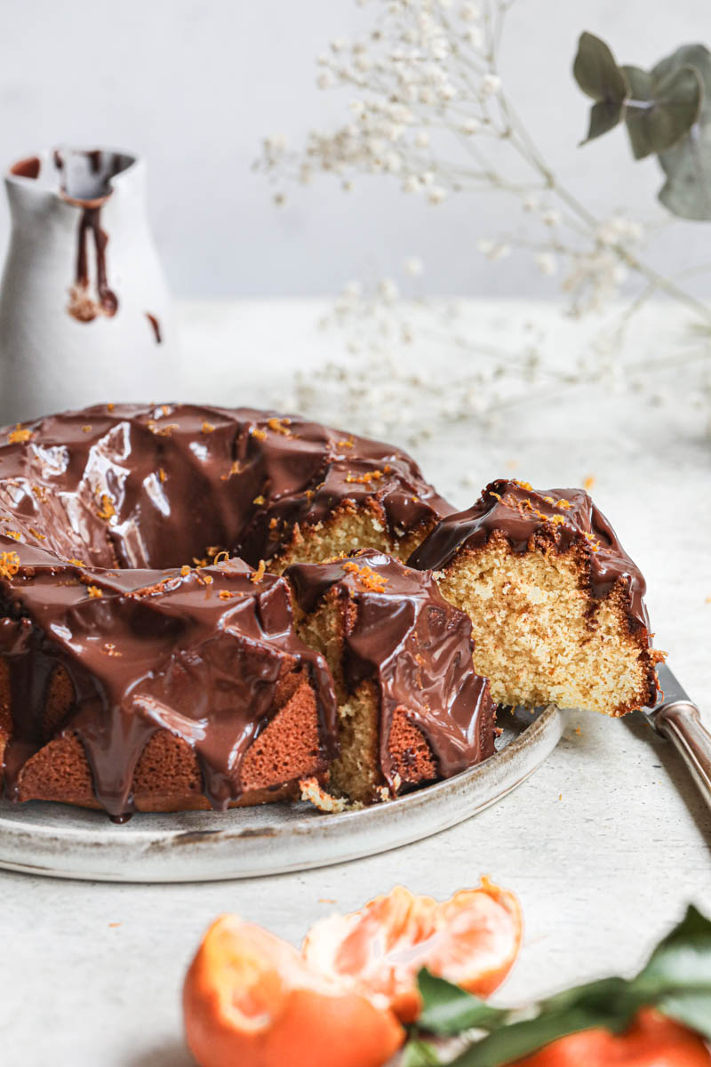 The glazed mandarin orange Bundt cake with one slice coming out on a grey plate with a knife on the side and a small grey jar in the back.