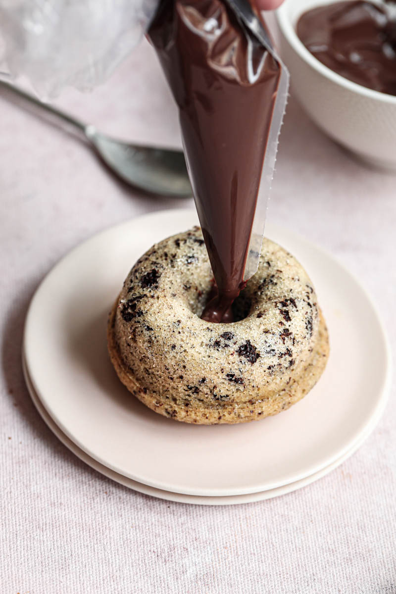 One hand piping the chocolate ganache onto one mini chocolate chip financier almond cake on a pink plate with a silver spoon on the side.