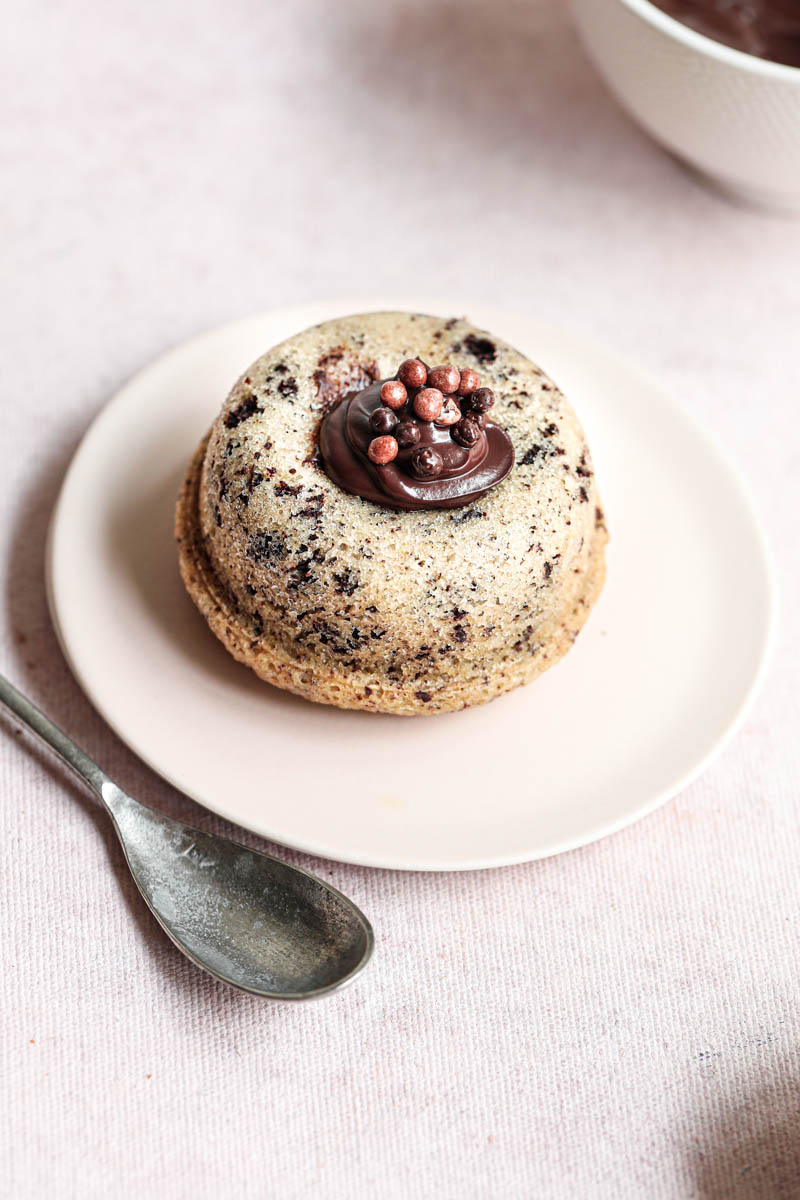 One mini chocolate chip financier almond cake with chocolate ganache on a pink plate with a silver spoon on the side and a bowl on the right top corner.