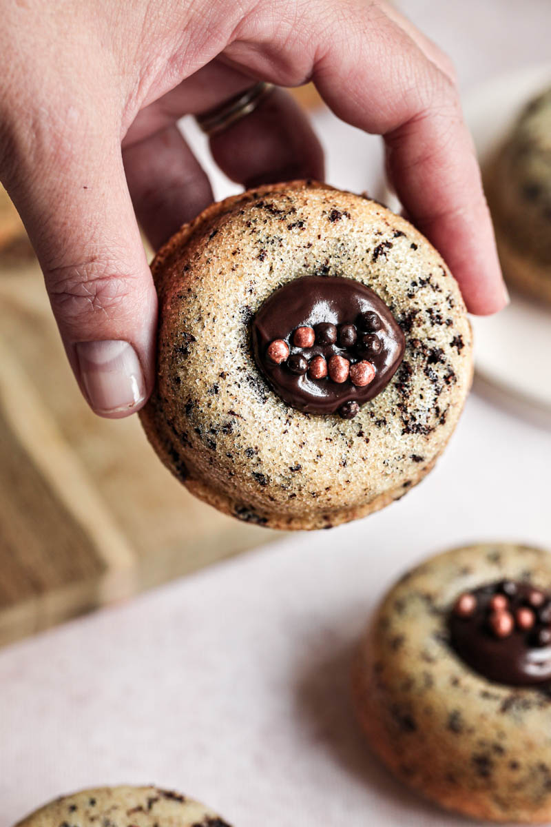 One hand holding one mini chocolate chip financier almond cake filled with chocolate ganache with others blurry in the back.