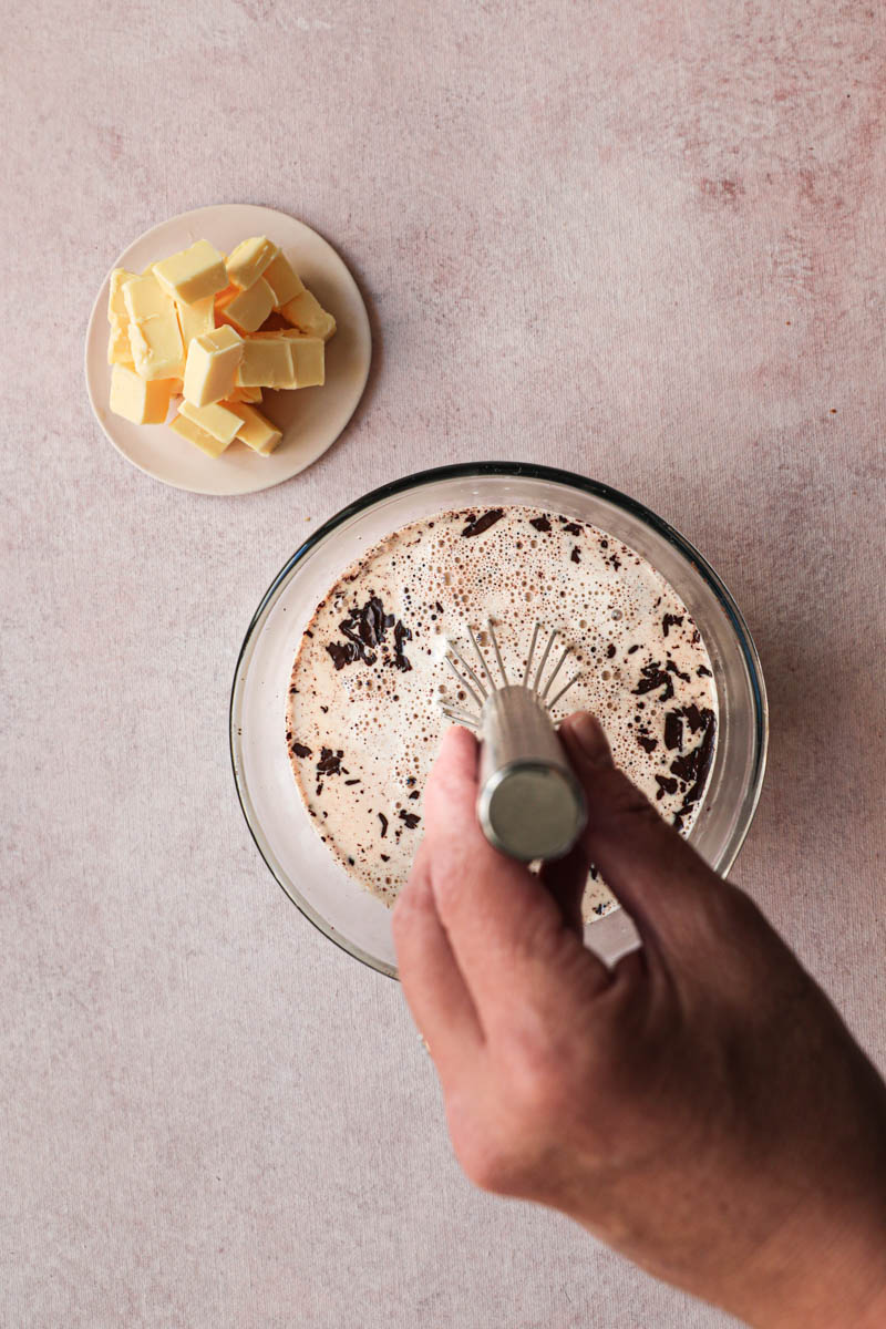 Chocolate ganache in the making: one hand whisking the chocolate and the hot heavy cream inside a glass bowl, with a small plate with butter on the side.