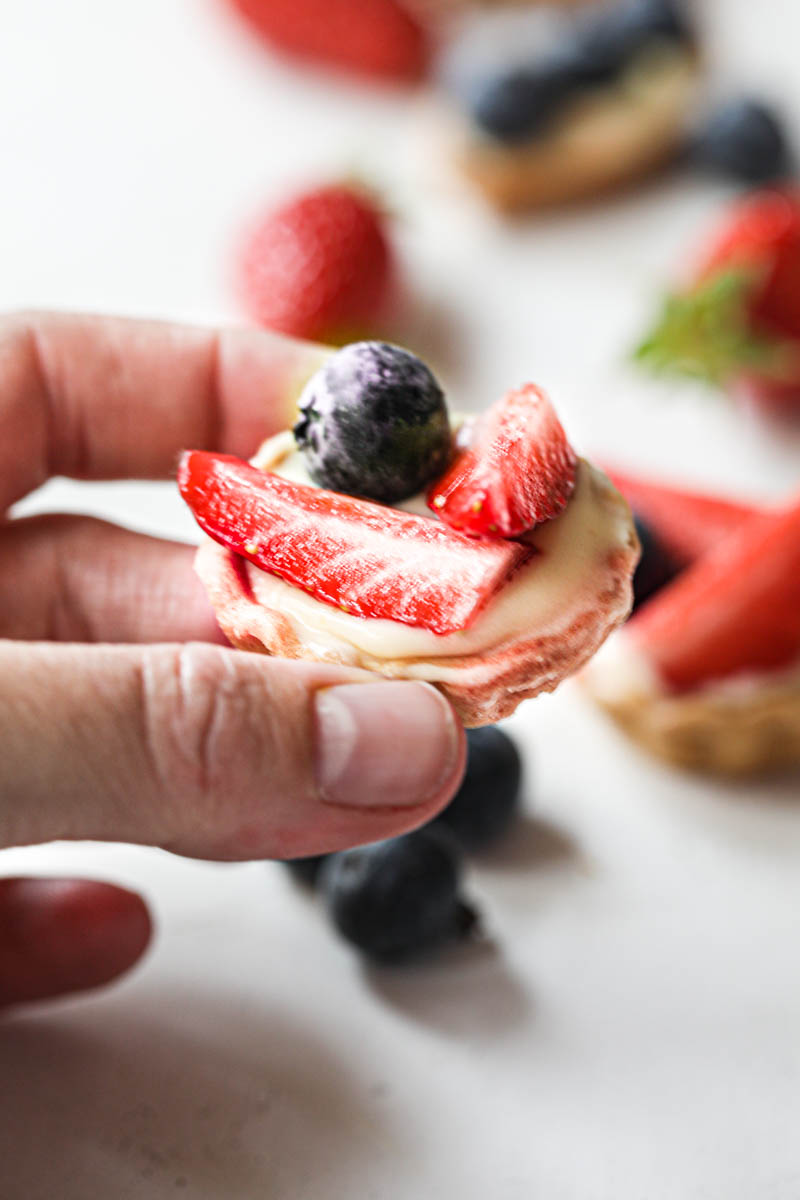 One hand holding a mini strawberry lemon tartlet with others blurry on the back of the frame.
