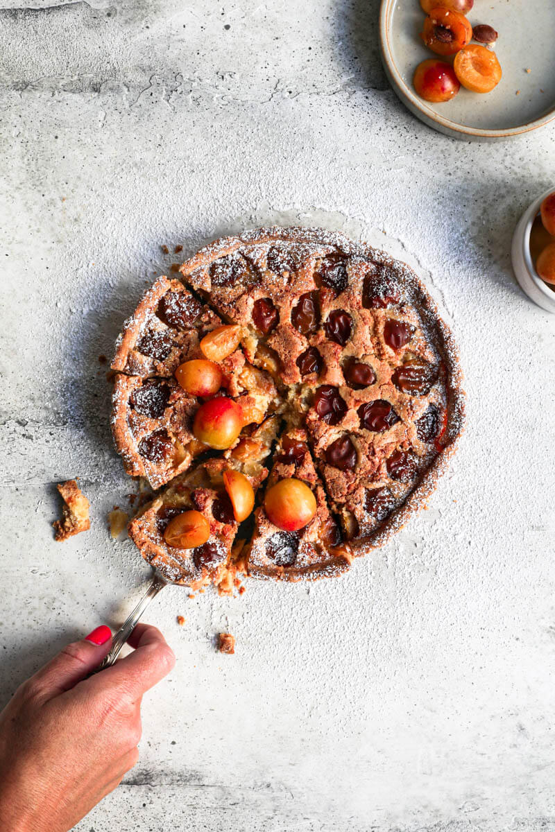 Overhead shot of the sliced Mirabelle plum tart with a hand holding the serving spoon