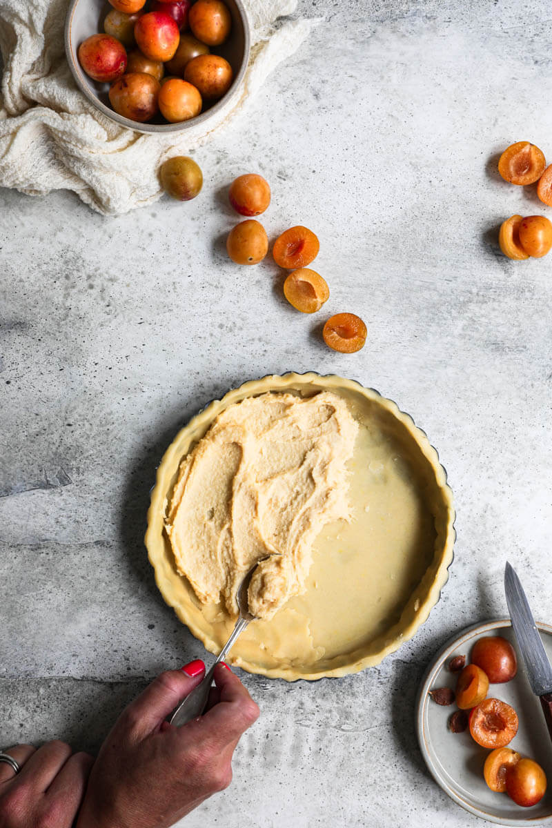 Overhead shot of the lined tart tin with a hand spreading the almond cream on the base