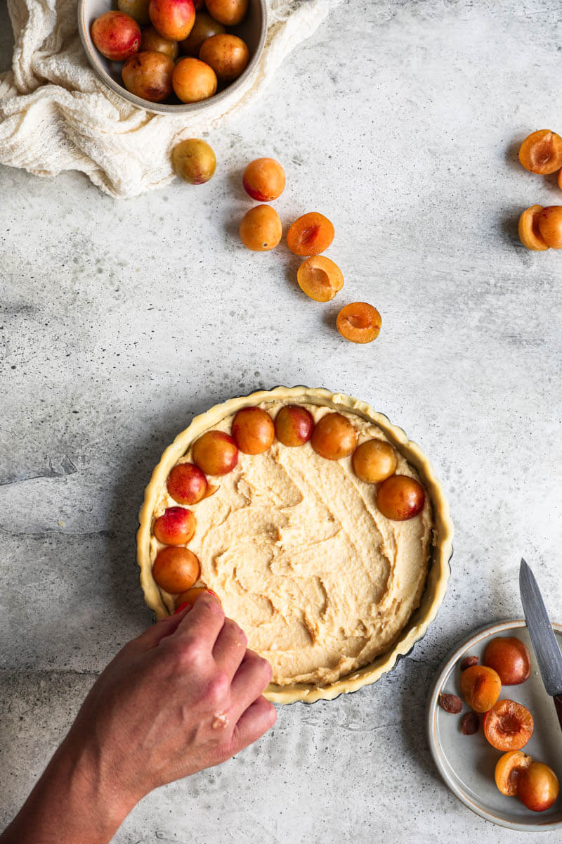 Overhead shot of the lined tart tin filled with almond cream with a hand placing the Mirabelle plums on top