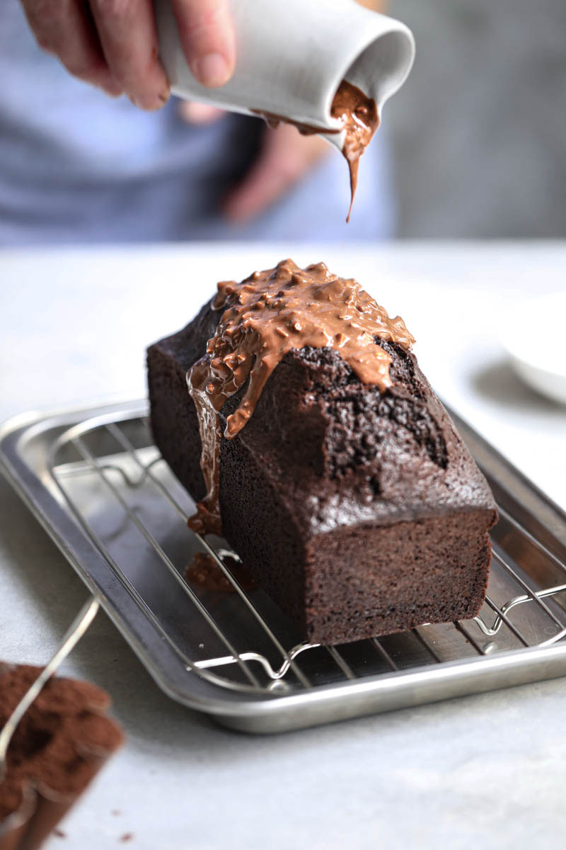 The baked one bowl chocolate cake on a wire rack with a hand pouring the almond chocolate glaze over it.