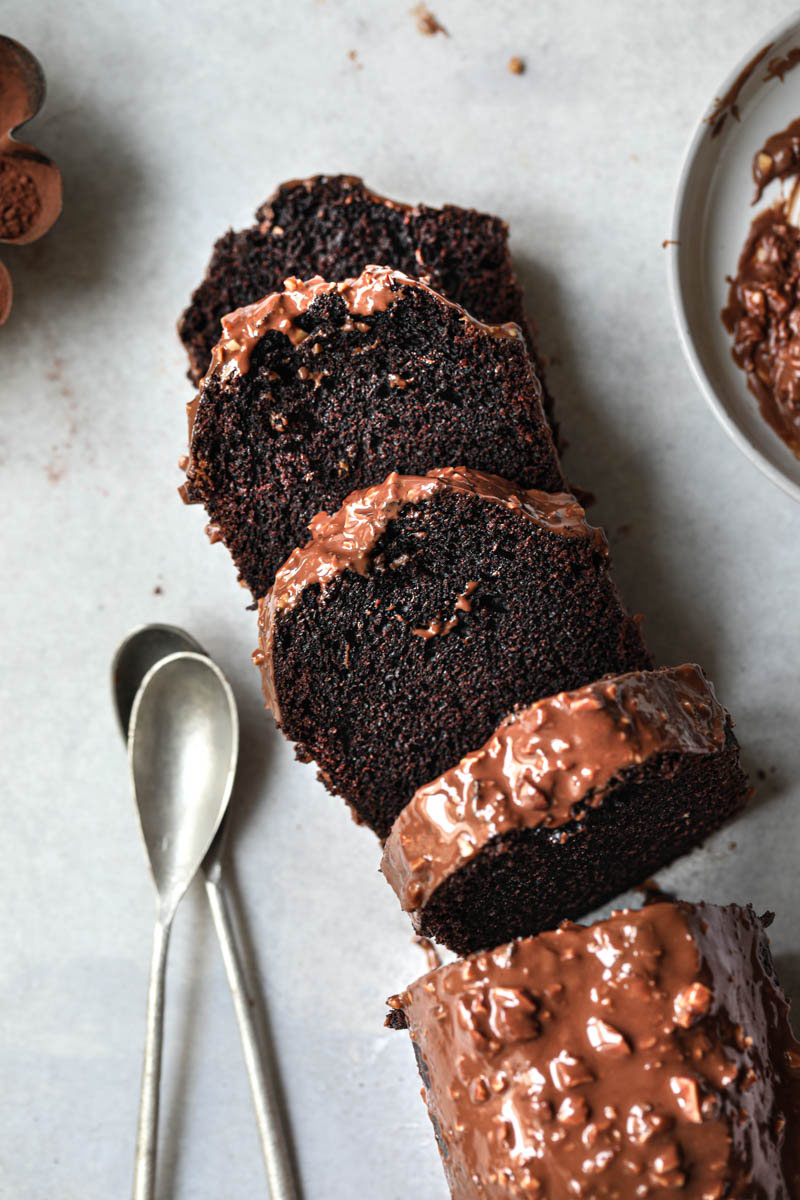 Sliced one bowl chocolate cake covered in almond chocolate glaze as seen from above with 2 spoons on the top side and a plate with extra glaze at the bottom.