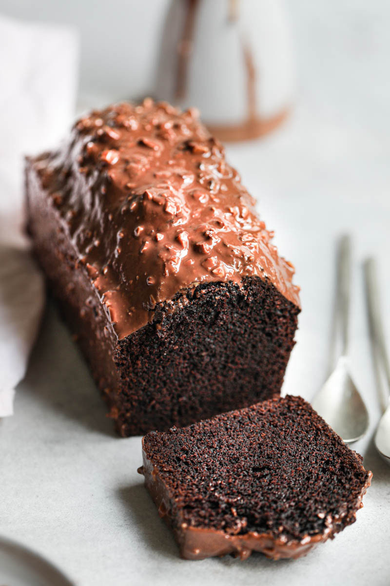 Whole one bowl chocolate cake covered in almond chocolate glaze with one slice cut and in the front of the photo, and a small ceramic jar in the back.