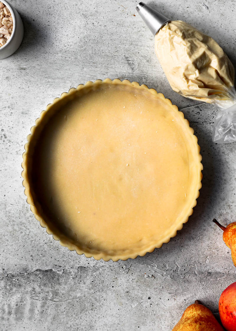 Overhead shot of the tart tin lined with the shortbread crust and the frangipane in a piping bag in the right corner