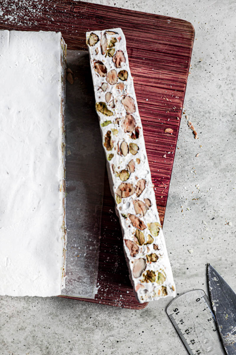 The nougat on a wooden cutting board, with one band cut off and facing up. A knife and a ruler can be seen in the bottom right corner.